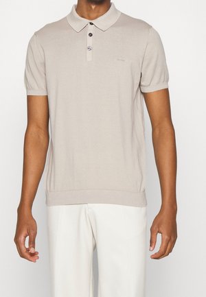 Man draagt een beige polo met korte mouwen en knoopsluiting en een witte broek, staand tegen een effen achtergrond.
