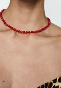 Collier ras du cou en perles rouges, composé de perles sphériques lisses et brillantes enfilées ensemble, porté sur un cou nu avec une tenue à imprimé léopard visible.