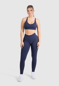 Granatowy stanik sportowy z głębokim dekoltem w połączeniu z legginsami o wysokim stanie. Dopasowany krój, gładka tekstura i bezszwowy design. Białe sneakersy.