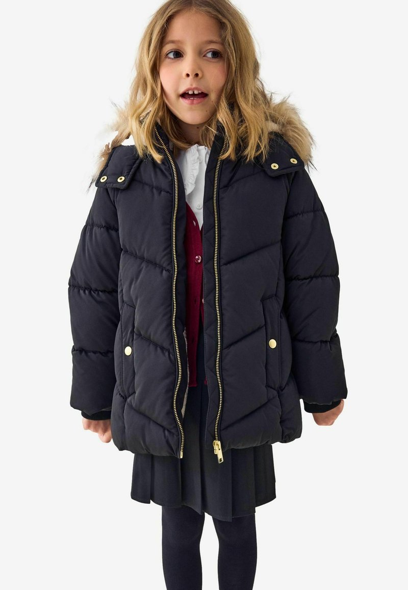Manteau matelassé noir avec capuche en fourrure, quincaillerie dorée et couture diagonale. Porté sur une blouse blanche et un pull rouge.
