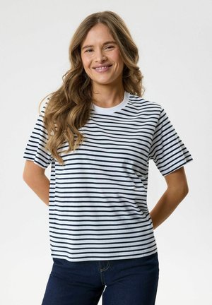 Jeune femme aux longs cheveux ondulés souriante, portant un t-shirt rayé horizontalement blanc et noir et un jean bleu foncé sur un fond uni.