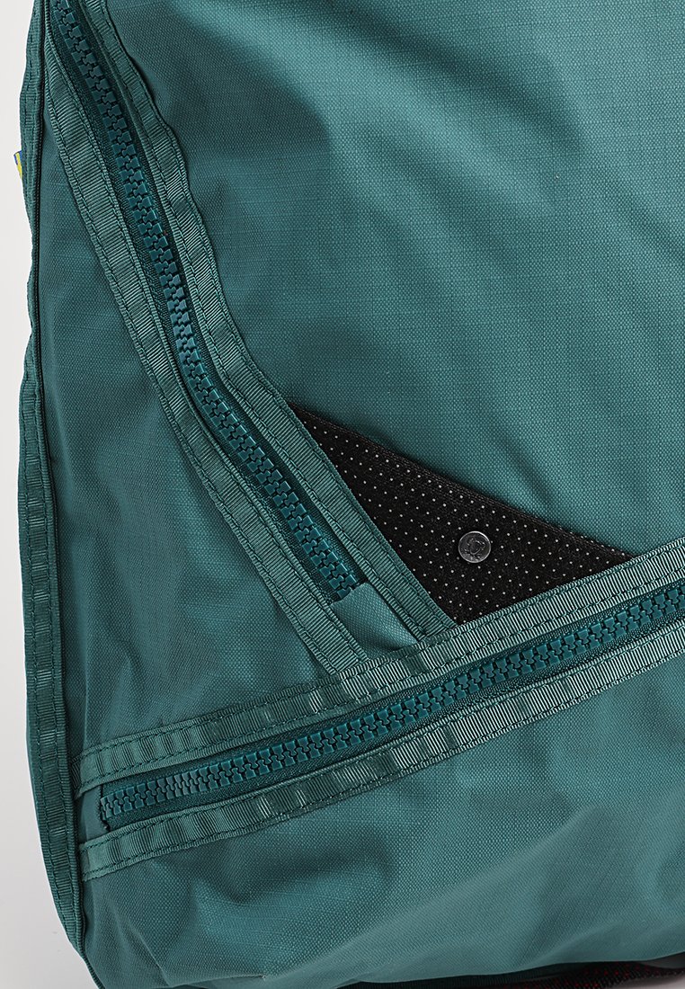 Türkiser Stoff-Rucksack mit zwei sichtbaren Reißverschlüssen und einer teilweise geöffneten Seitentasche, die einen schwarzen strukturierten Gegenstand im Inneren zeigt.