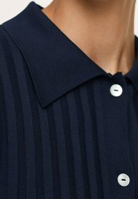 Camisa de cuello azul marino con textura de canalé vertical y cinco botones blancos, con franjas contrastantes para aportar interés visual.