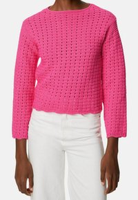 Pull rose en tricot crochet avec manches longues et ourlet festonné. Présente un motif en grille et une texture respirante. Porté avec un pantalon blanc.