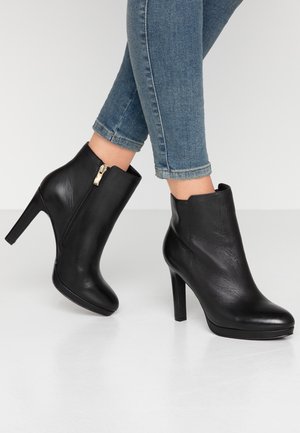 Person iført slanke blå jeans og sorte læderankelstøvler med sidelås og høje hæle, stående på hvidt gulv.
