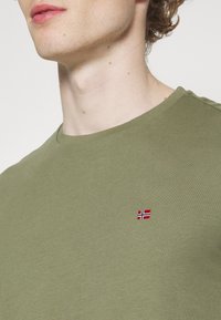 Oliven grøn t-shirt i bomuld med rund udskæring og et lille broderet norsk flag på venstre bryst. Glat tekstur.