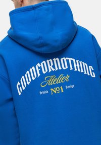 Felpa blu con una grande grafica testuale bianca e gialla sul retro, che presenta "GOODFORNOTHING Atelier No.1 British Design". Tessuto morbido.