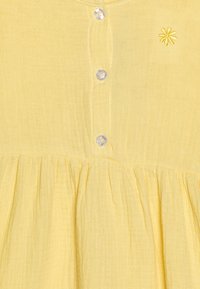 Gelbes Baumwollkleid mit strukturiertem Stoff, rundem Ausschnitt und fünf Knopfverschlüssen mit einem kleinen bestickten Blumenmotiv.