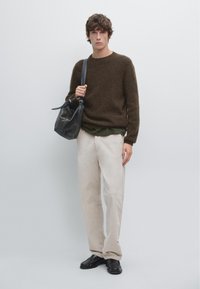 Un pull en laine marron, un sous-vêtement vert, un pantalon beige et des chaussures noires. Tenant un sac en cuir noir au design décontracté. Look simple et moderne.