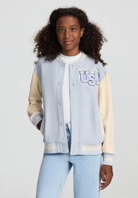 Jasnoniebieska i kremowa kurtka varsity z zatrzaskami, ściągaczami przy rękawach i wyraźną naszywką "USA" na klatce piersiowej. Miękka tekstura materiału.