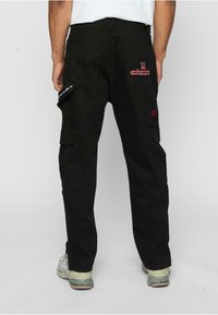 Svarte cargo-bukser med flere lommer, broderte detaljer og løs passform. Stoffet virker slitesterkt med en glatt tekstur.