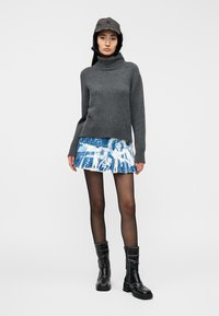 Γκρι πλεκτός πουλόβερ με ψηλό λαιμό, πλισέ φούστα με μπλε και λευκό μοτίβο, διαφανή μαύρα καλσόν και μαύρα μποτάκια αστραγάλου με λεία επιφάνεια.