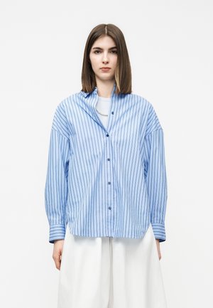 Mladá žena s rovnými hnedými vlasmi, oblečená v voľnej svetlomodrej košeli s prúžkami a bielych širokých nohaviciach na bielom pozadí.