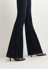 Höga, mörkblå utsvängda jeans med en slät textur. Tillsammans med svarta stilettklackar med tunna remmar och öppen tå.