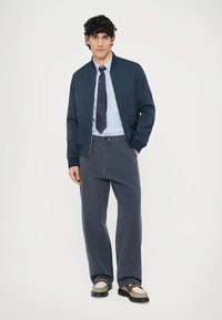 Jūras kara aviācijas bomber jaka, gaiši zila krekla krekliņš, rakstaina kaklasaite, pelēkas platu kāju bikses un divtoņu apavi. Minimālistisks dizains un neitrālas krāsas.