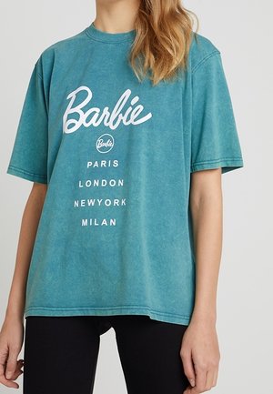 Vrouw draagt een turquoise oversized Barbie T-shirt met de namen Parijs, Londen, New York en Milaan in witte letters, gecombineerd met een zwarte broek.