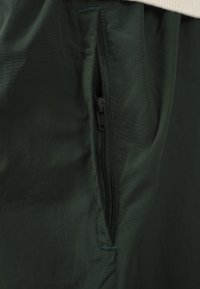 Gros plan d'une poche de vêtement vert foncé avec une fermeture éclair noire partiellement fermée et des détails de couture visibles sur le tissu.