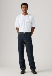 Camiseta blanca de manga corta con un bolsillo en el pecho, combinada con unos pantalones vaqueros de pierna ancha de mezclilla azul oscuro que presentan costuras en contraste y un ajuste holgado.