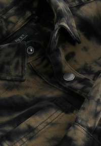 Giacca in corduroy tie-dye nera e marrone con colletto, chiusura a bottoni e dettagli di cucitura visibili. L'etichetta reca la scritta "NEXT."
