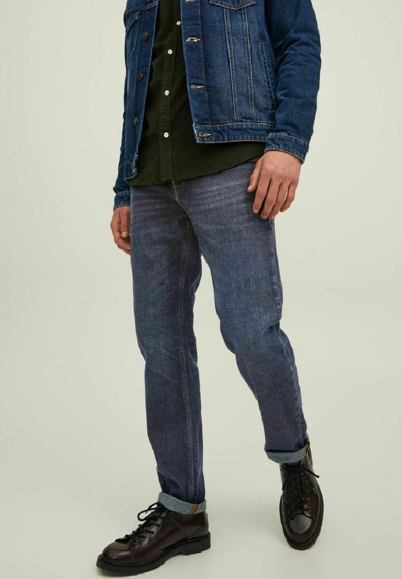 Vaqueros de mezclilla azul oscuro con corte recto, puños enrollados de color azul claro, combinados con una camisa verde y botas negras de cordones.