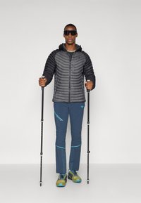 Veste noire rembourrée avec une capuche, pantalon de sport bleu et bâtons de randonnée. Surface texturée sur la veste ; accents bleus sur le pantalon et les chaussures.