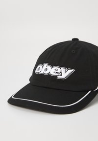 Fekete sapka ívelt csúccsal, amelyen fehér hímzett "obey" logó és fehér díszítés található a széleken. Pamut anyagból készült.
