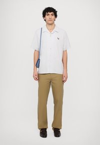 Striped short-sleeve shirt in white and light blue, buttoned with a small colored emblem. Paired with tan pants and brown shoes.