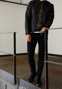 Schwarze Lederjacke, gemusterter Pullover in Dunkelgrün und Gelb, schwarze Slim-Fit-Jeans und schwarze Schnürschuhe. Steht auf einem Metallgeländer.