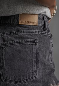 Shorts en denim noir avec une poche arrière et une étiquette en cuir beige embossée avec "SUPERDRY". Le tissu a une texture subtile.