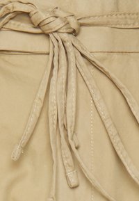 Tecido bege com detalhe de cinto amarrado. A textura é suave com costuras visíveis. Laços de tecido longos e finos pendem para baixo.