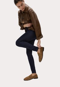 Mocasines de ante marrón con punta redonda, que presentan un detalle fruncido en la parte delantera. Combinados con jeans ajustados oscuros y una chaqueta de cuero marrón.
