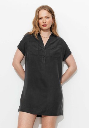 Jeune femme aux cheveux blonds ondulés portant une robe-chemise noire à manches courtes avec des poches poitrine, debout devant un fond blanc.