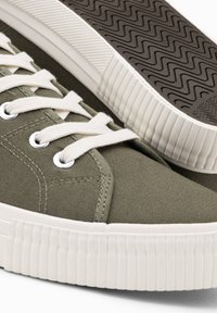 Olivgrüne Segeltuch-Sneaker mit weißen Gummisohlen, runden Schnürsenkeln, Metallösen und strukturiertem Muster auf der Außensohle.