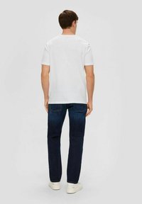 Weißes Baumwoll-T-Shirt mit kurzen Ärmeln, von hinten betrachtet, kombiniert mit dunkelblauen Jeans und weißen Sneakern, vor einem neutralen Hintergrund.