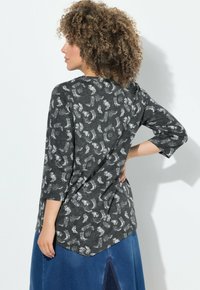 Mujer con cabello rizado que lleva una blusa de manga ¾ de color gris oscuro con estampado y una falda de mezclilla azul, de pie con la mano en la cadera, mirando hacia un lado.