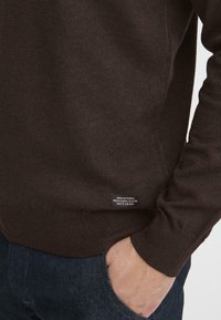Brun strikkegenser med ribbestruktur, med et lite merke ved sømmen. Båret med mørkeblå denimjeans.