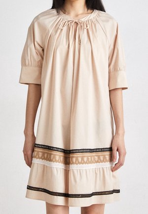 Femme portant une robe beige ample avec des manches aux coudes, un col à nouer et une bordure décorative noire, marron et blanche près de l'ourlet.