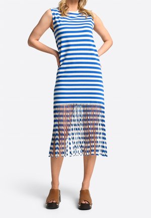 Femme portant une robe sans manches à rayures bleues et blanches avec un ourlet à franges, associée à des sandales à plateforme marron à bout ouvert, debout les mains sur les hanches.