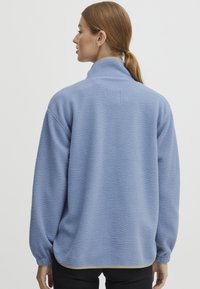 Ljusblå fleece-pullover med hög krage, texturerad tyg, avslappnad passform och långa ärmar. Har en böjd nederkant och minimal sömnad.