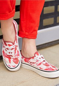 Baskets en toile rouges et blanches avec un motif camouflage, lacets blancs et semelles en caoutchouc noires, portées avec un pantalon rouge retroussé.