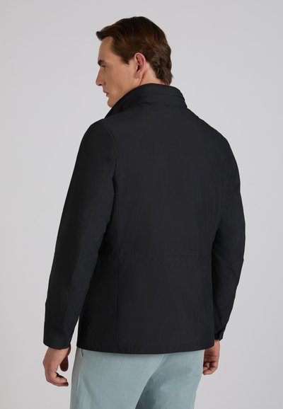 Mężczyzna zwrócony bokiem, ubrany w czarną kurtkę z wysokim kołnierzem i jasnoniebieskie spodnie, na tle jednolitego szarego tła.