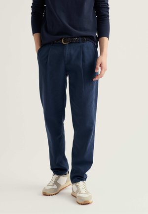 Persoon gekleed in donkerblauwe geplooide broek met zwarte gevlochten riem, witte en beige sneakers, en donkerblauw shirt met lange mouwen, staand met handen in de zakken.