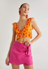 Top corto naranja con patrón floral, mangas con volantes y un lazo en la parte delantera; combinado con una falda envolvente rosa con un lazo y herrajes dorados.