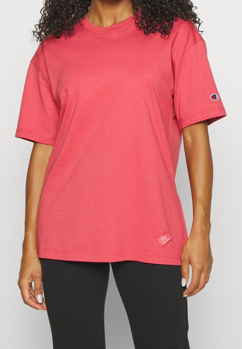 Femme portant un t-shirt rouge ample à manches courtes avec un petit logo sur la manche gauche et un pantalon noir sur fond uni.