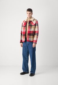 Checked wool jacket in red, black, and beige, with a soft collar and buttons; paired with blue denim jeans and black shoes.