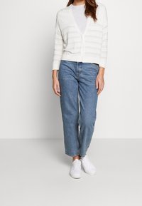 Vit knapp-up cardigan med vertikala ränder, bärs över en ljusgrå skjorta. Kombineras med lösa blå jeans och vita sneakers.