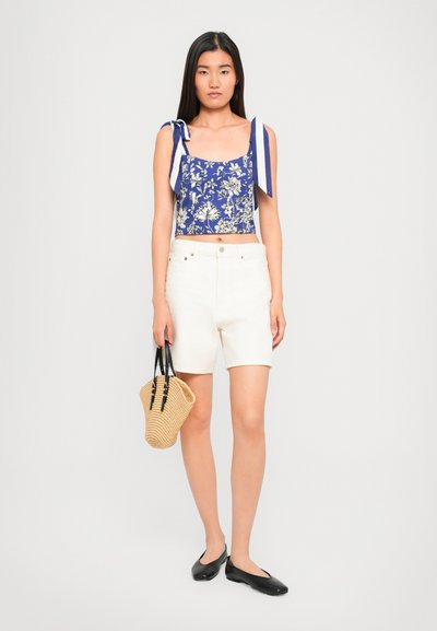 Woman wearing blue floral crop top with tied shoulder straps, white shorts, black flats, holding tan woven handbag against white background.
