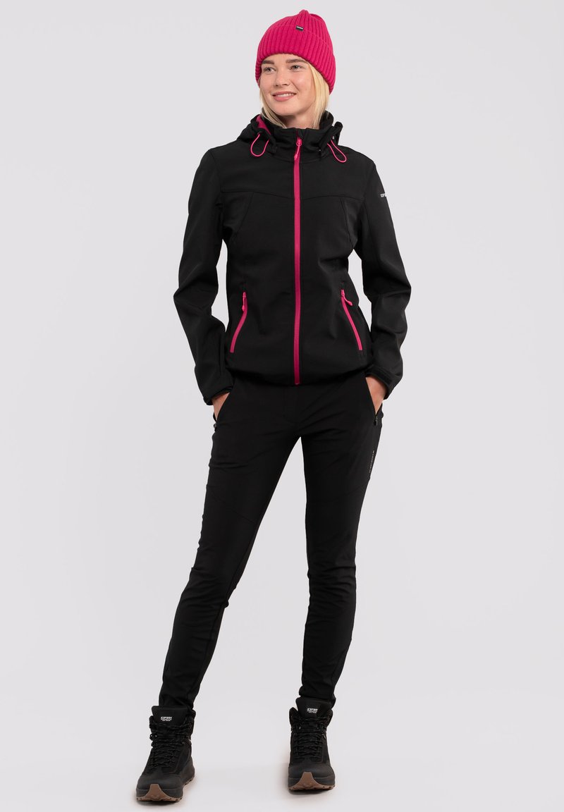 Giacca softshell nera con zip e dettagli rosa, abbinata a pantaloni neri e scarponi da trekking neri. Il modello indossa un berretto rosa.