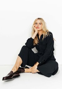 Combinaison noire à boutons avec des manches longues et des jambes larges. Assortie à des mocassins marron foncé avec des clous. Bracelet jonc doré au poignet.