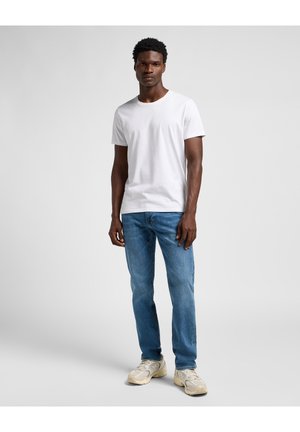 White crewneck t-shirt paired with light blue straight-leg jeans and beige sneakers. Model stands against a plain light background.